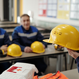 Hombre sostiene un casco amarillo en primer plano, con otros hombres sentados al fondo en un aula